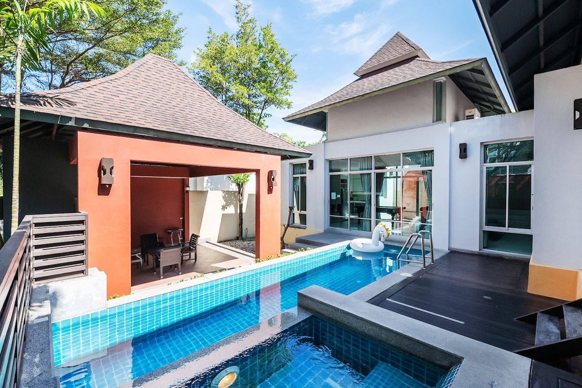 A private pool villa is depicted with a clear blue swimming pool at its center, flanked by modern architecture. Natural light floods a nearby patio area, which features seating for relaxation. Lush greenery surrounds the property, enhancing the tranquil atmosphere.