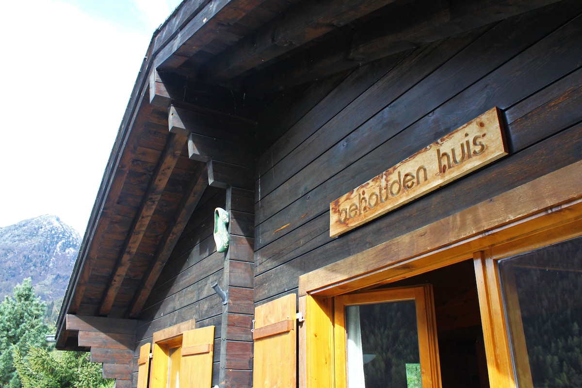 The exterior of the chalet features natural wooden siding with a rustic charm. A wooden sign with the name 'Behouden Huis' is prominently displayed above the entrance. Large windows allow for abundant natural light, and the surrounding greenery enhances the inviting setting.