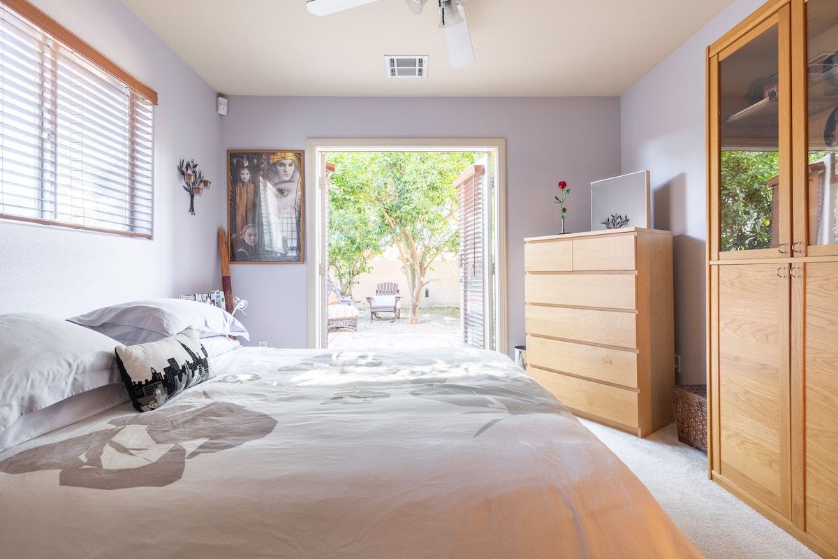 A spacious bedroom is illuminated by natural light, featuring a comfortable bed with a neatly arranged white duvet. A wooden dresser stands against the wall, while a sliding glass door leads to a private patio area surrounded by greenery.