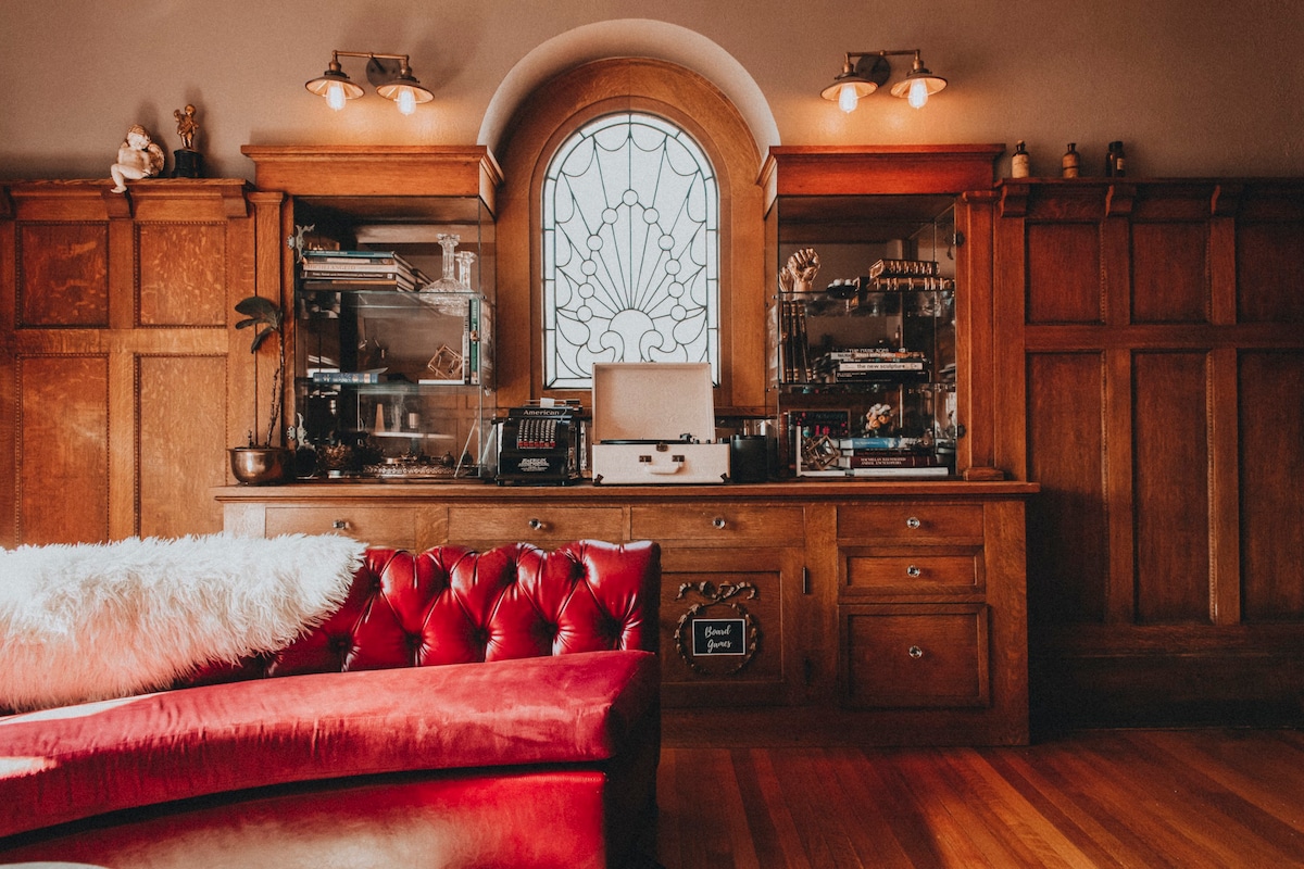 A vintage wooden sideboard is displayed under an arched stained glass window, adding historical charm. A plush red couch invites relaxation, accompanied by decorative items on the shelves above. Warm lighting highlights the room's textures and wood finishes, creating a cozy atmosphere.