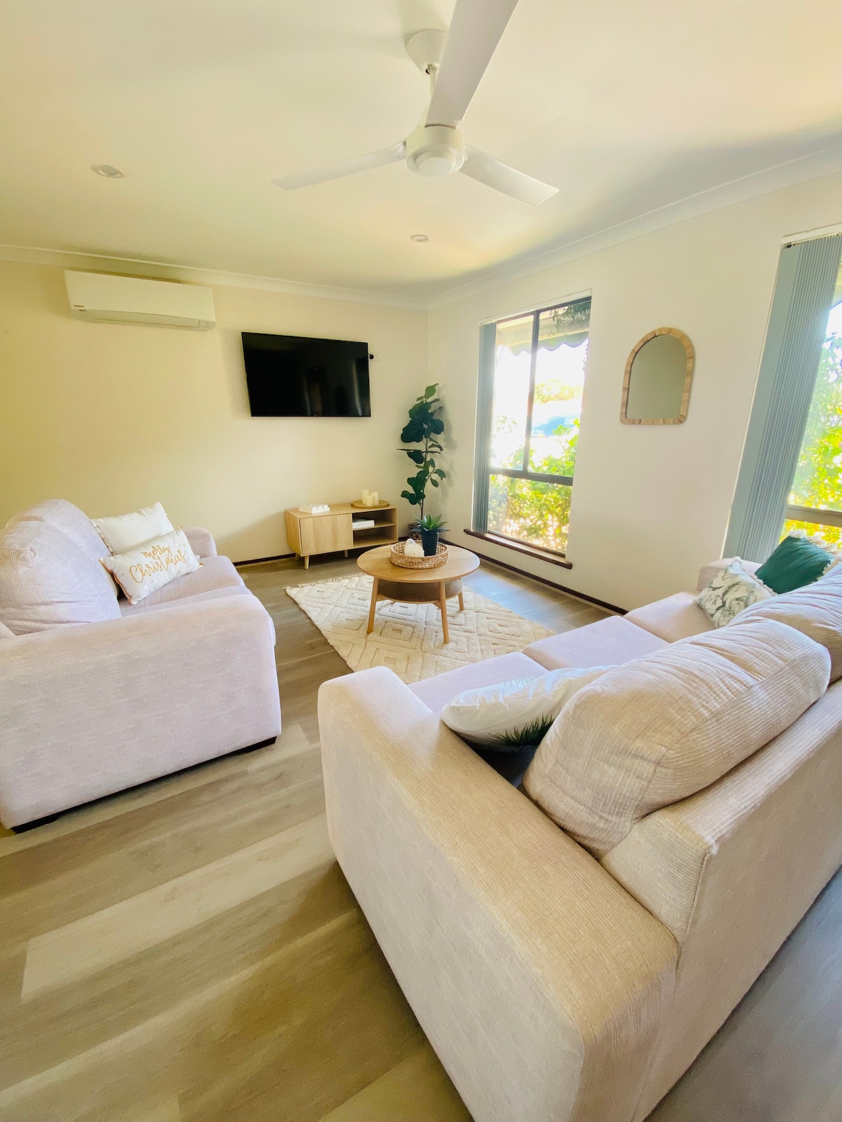 A cozy lounge area features two light-colored sofas facing a round wooden coffee table. A flat-screen TV is mounted on the wall, and large windows provide natural light, framed by soft curtains. A potted plant adds a touch of greenery to the space.