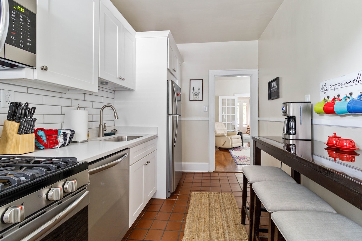A modern kitchen features sleek white cabinetry and stainless-steel appliances. A gas range is positioned adjacent to a refrigerator, while a small dining table with three cushioned stools is situated against a wall. A neutral rug enhances the warm tile flooring.
