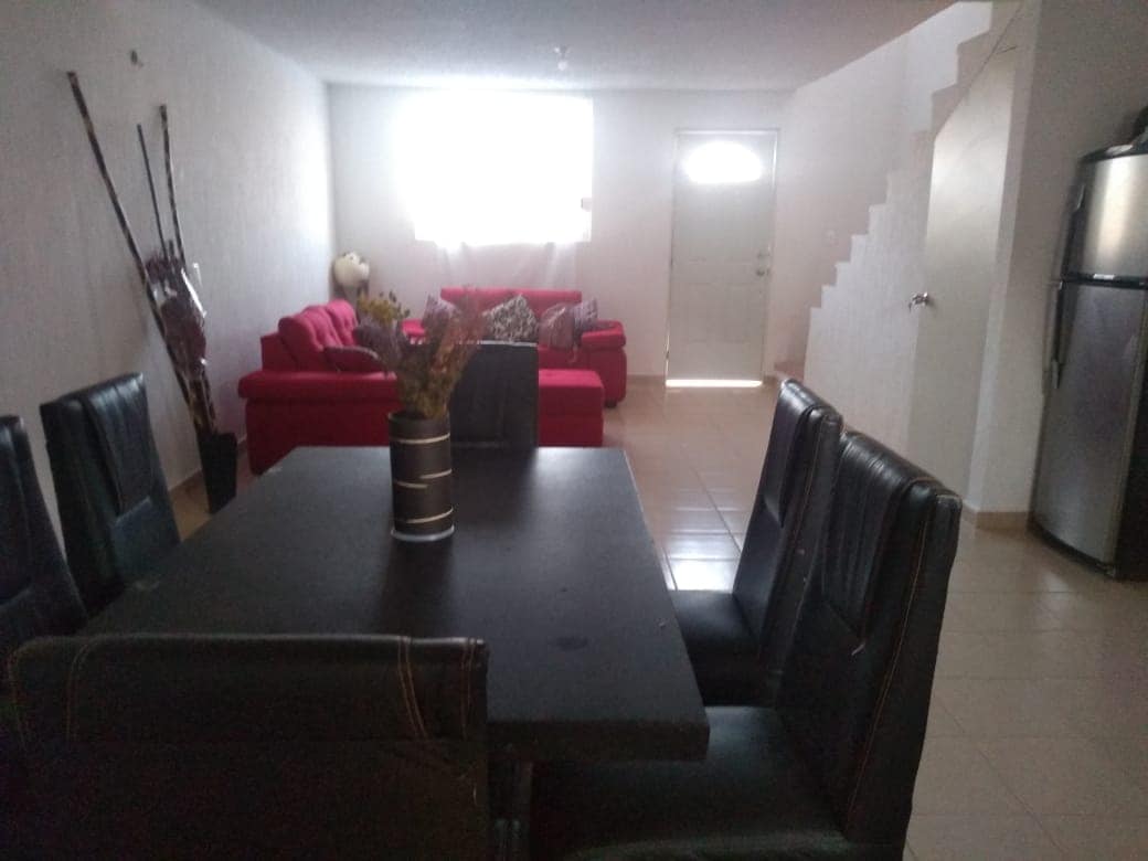 A dining area features a dark table surrounded by six upholstered chairs. A vase with dried flowers is centered on the table. In the background, a red sectional sofa is positioned next to a door and a staircase, with natural light illuminating the room.
