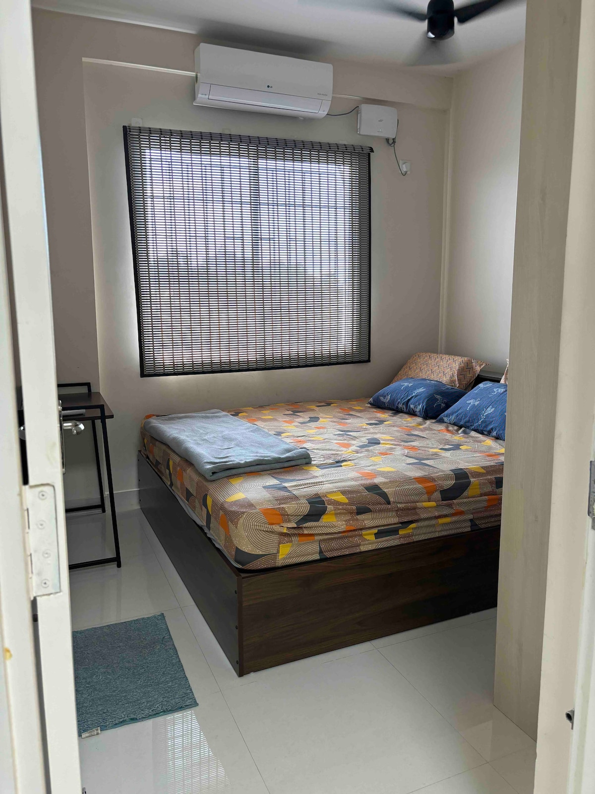 A cozy bedroom features a spacious bed with patterned bedding, accompanied by two decorative pillows. An air conditioning unit is mounted above a window with a black-out blind. A small bedside table is positioned next to the bed, and a soft rug lies on the floor.
