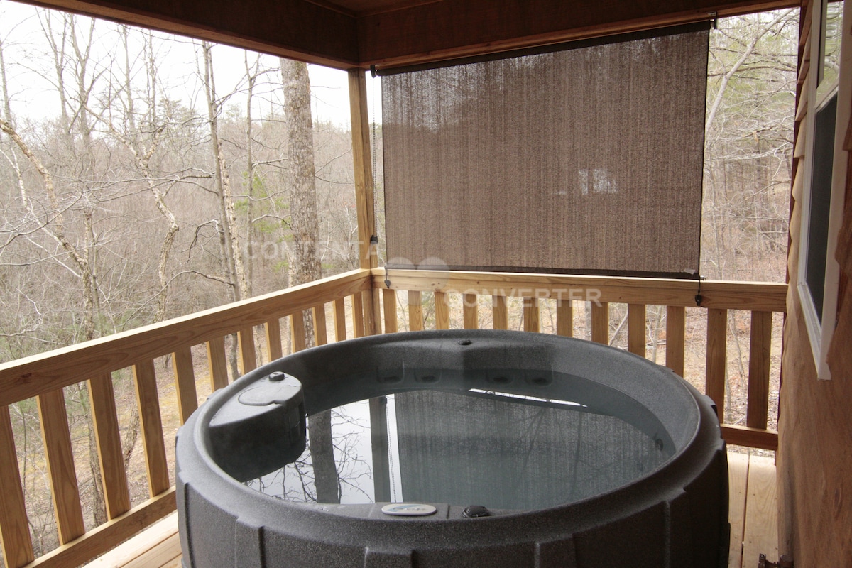 A hot tub is positioned on the back deck of the cabin, surrounded by natural privacy from nearby trees. The tub features curved edges and built-in seating, inviting relaxation. A half-closed privacy screen offers a sense of seclusion while enjoying the view of the surrounding woodland.