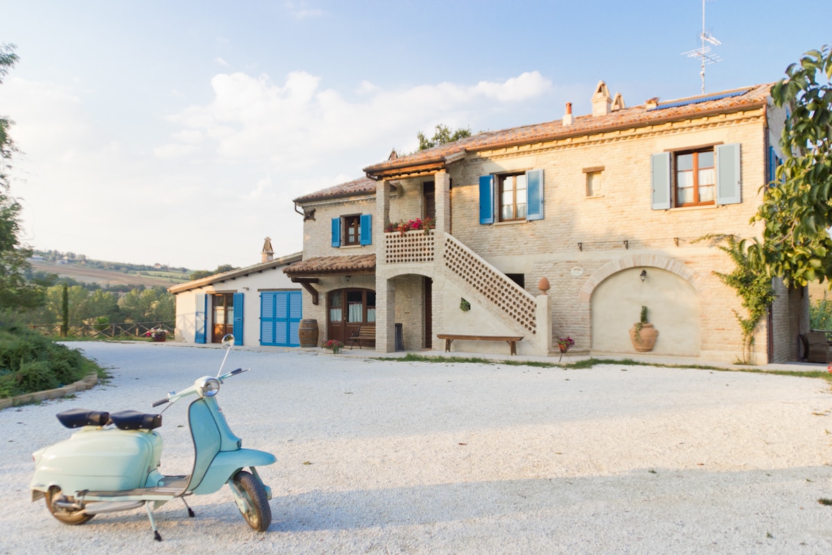 A charming farmhouse with a rustic facade is set in a serene landscape. Light blue shutters adorn the windows, and a staircase leads to an upper level. A vintage scooter is positioned in the foreground, framed by gravel and greenery.