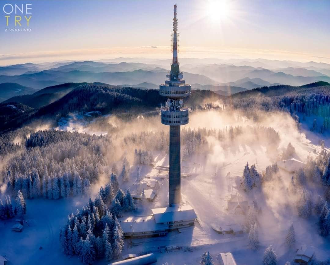 A tall broadcasting tower rises above a blanket of snow, surrounded by dense evergreen trees. Mist gently rolls through the winter landscape, with distant mountains visible under a clear sky illuminated by sunlight.