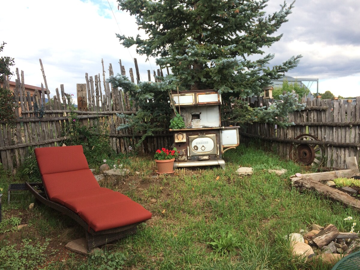 A lounge chair with deep red cushions rests on a grassy area in the private garden. An antique stove stands nearby, surrounded by greenery and rustic wooden fencing that encloses the space.