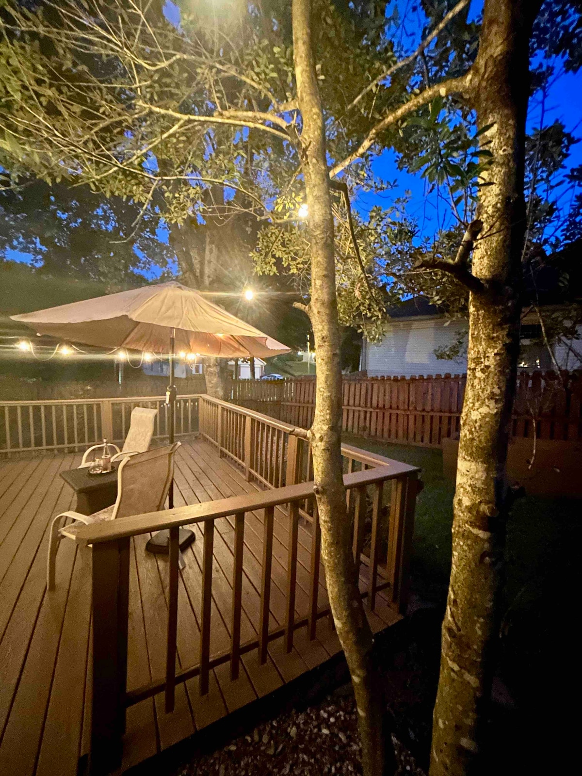 A peaceful deck area is seen during twilight, featuring an outdoor umbrella and two chairs. Warm cafe string lights create a cozy ambiance while the fenced yard provides privacy. Trees add natural elements to the serene outdoor setting.