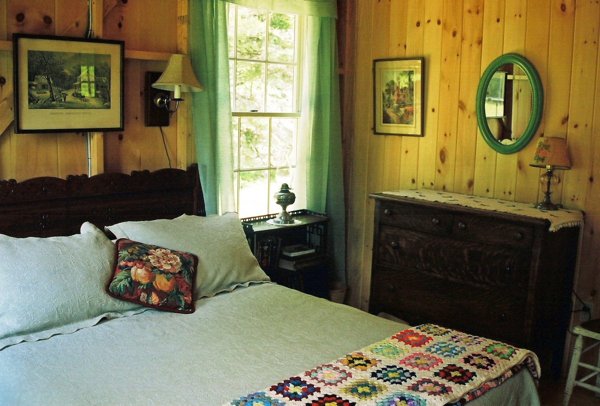 A warm and inviting bedroom features a spacious bed dressed in light-colored linens. Wooden paneling lines the walls, complementing the vintage dresser and framed artwork. Natural light filters through the window dressed with sheer curtains, while a colorful quilt adds a pop of color at the foot of the bed.