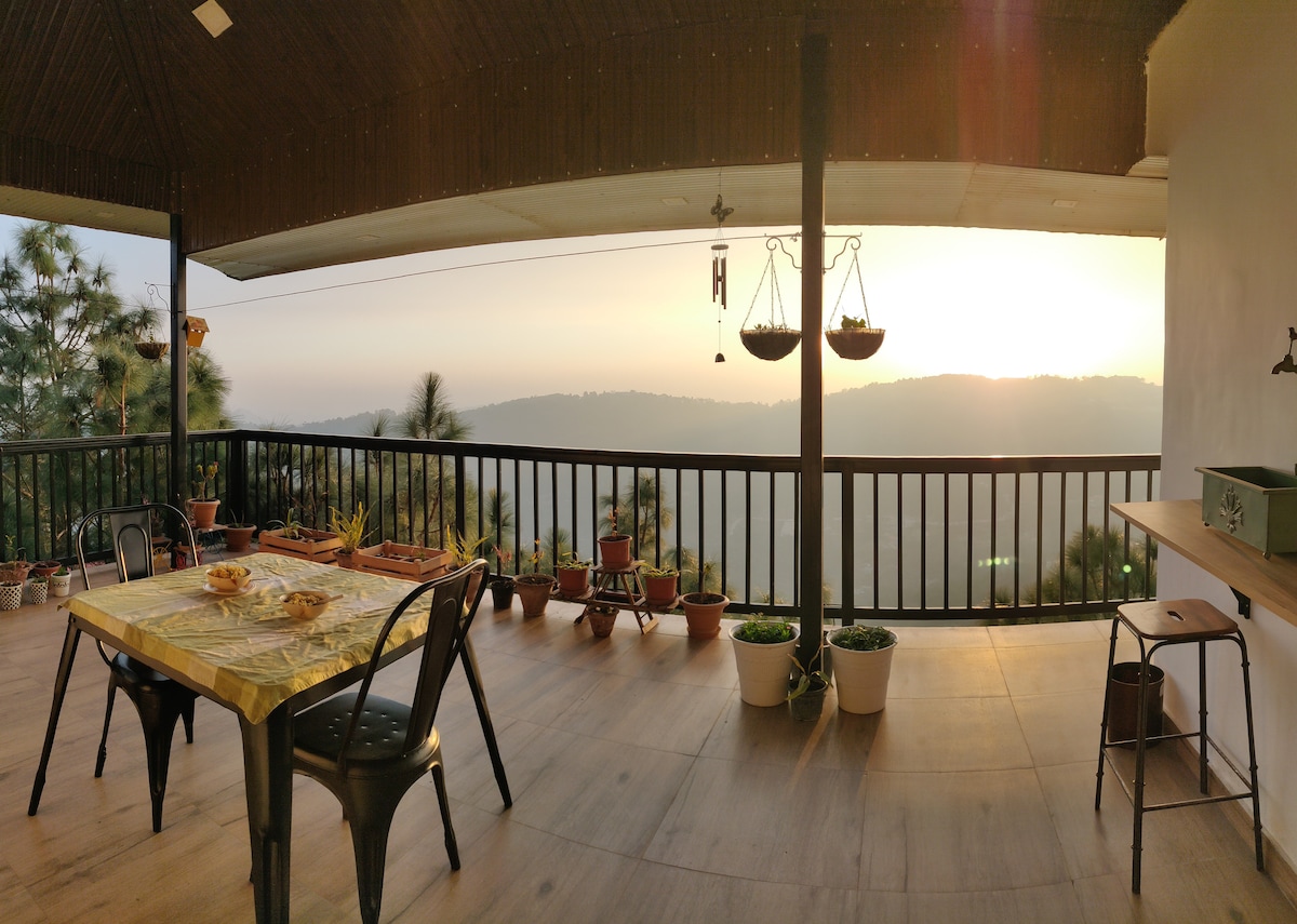 An outdoor dining area features a table set for meals, surrounded by chairs. Hanging planters add greenery, while pots line the railing. A scenic view of the distant hills is visible, illuminated by soft morning light, creating a serene atmosphere.