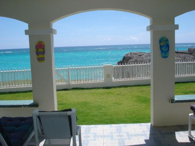 A view from the covered porch displays vibrant turquoise waters meeting a clear blue sky, framed by white arches. The grassy area extends toward a white fence, highlighting the tranquility of the oceanfront setting.