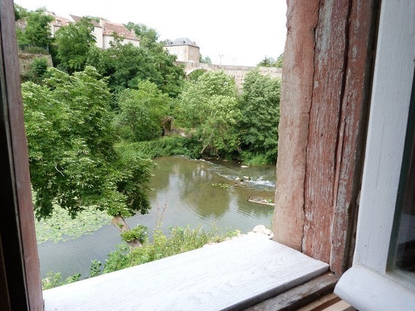 A view from a window showcases the tranquil river Armençon, surrounded by lush greenery and trees. The water gently flows past, reflecting the vibrant colors of nature. A bridge and historic buildings can be faintly seen in the distance, enhancing the serene atmosphere.
