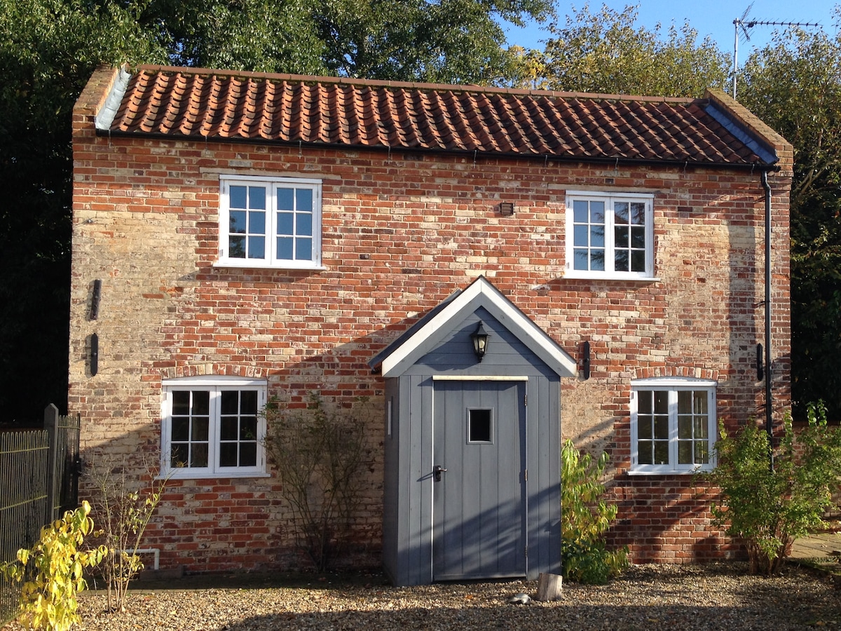 A rustic cottage with a brick facade features a central grey door flanked by two sets of white-framed windows. The roof showcases traditional red tiles, while surrounding greenery complements the structure. The setting conveys a sense of charm and tranquility.