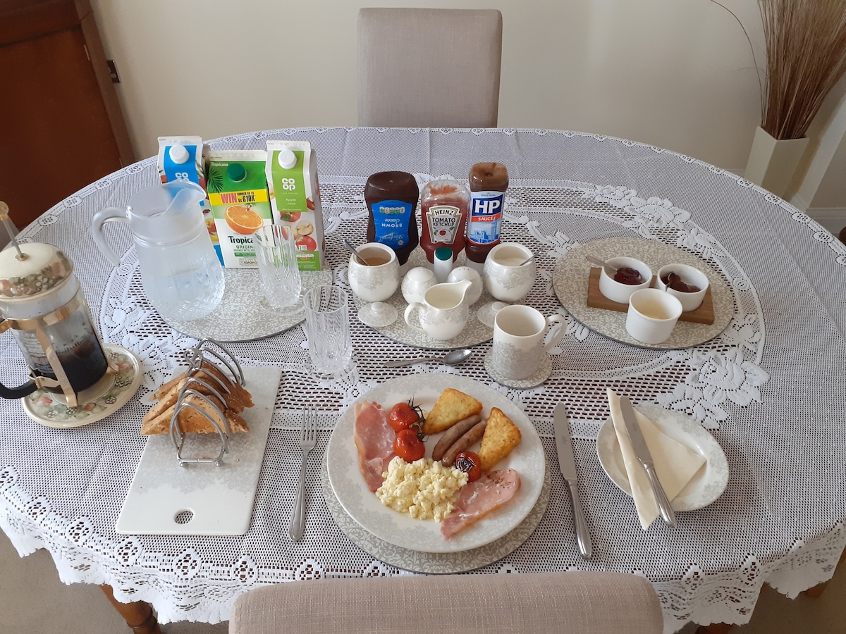 A dining table is set with a variety of breakfast items, including plates of eggs, bacon, tomatoes, and toast. Condiments, juices, and beverages are neatly arranged in containers. The table is covered with a lace tablecloth, and dinnerware is presented in a simple, elegant style.