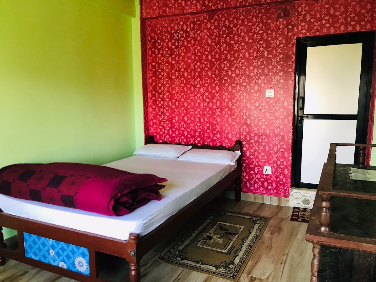 A bright room is adorned with contrasting green and red walls, featuring a spacious bed with a red blanket layered on top. A small table is placed beside the bed, and natural light enters through the nearby window with a black frame.