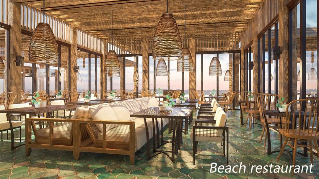 An airy beach restaurant features large windows providing expansive views. The space is adorned with wooden furnishings and woven pendant lights. Tables are set with elegant tableware, and a neutral-toned sofa is positioned against one wall, enhancing the welcoming dining atmosphere.