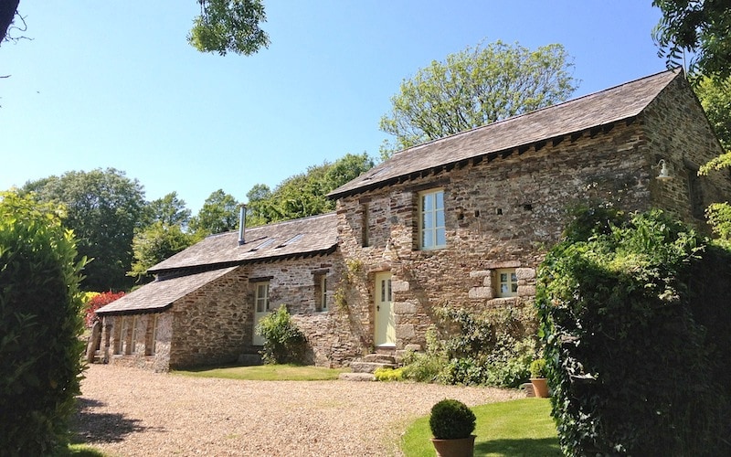 The exterior of Long House showcases a charming stone façade with large windows, allowing natural light to fill the interior. A gravel pathway leads to the entrance, framed by lush greenery and well-manicured hedges, creating a welcoming ambiance.