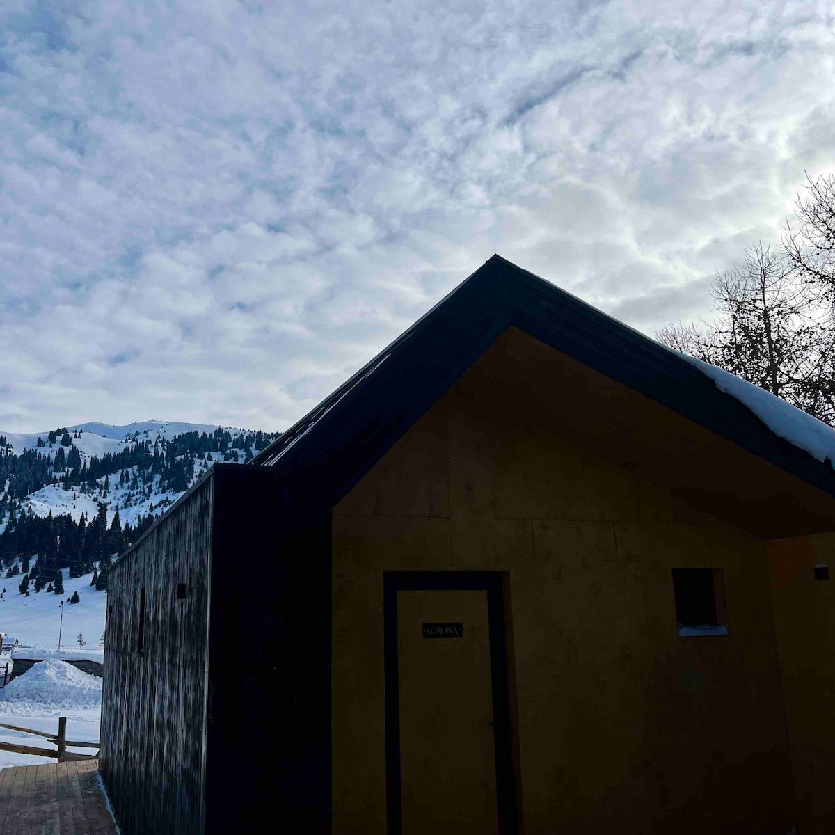 The exterior of a cottage is shown against a backdrop of a cloudy sky and snow-covered mountains. The building features a minimalist design with a sloped roof and a single door visible. Snow blankets the ground around the structure.