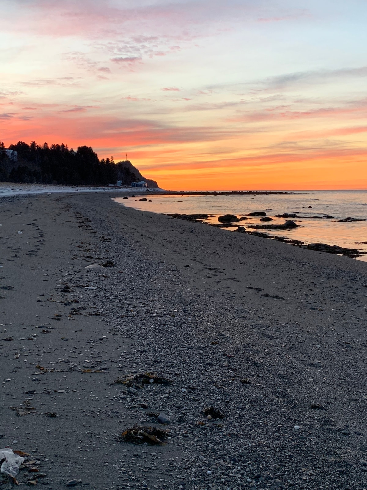 The beach is lined with smooth sand and scattered stones, reflecting the soft colors of the sunset. Gentle waves lap at the shore, while the sky is painted in warm hues of orange and pink, with silhouettes of trees visible in the background.