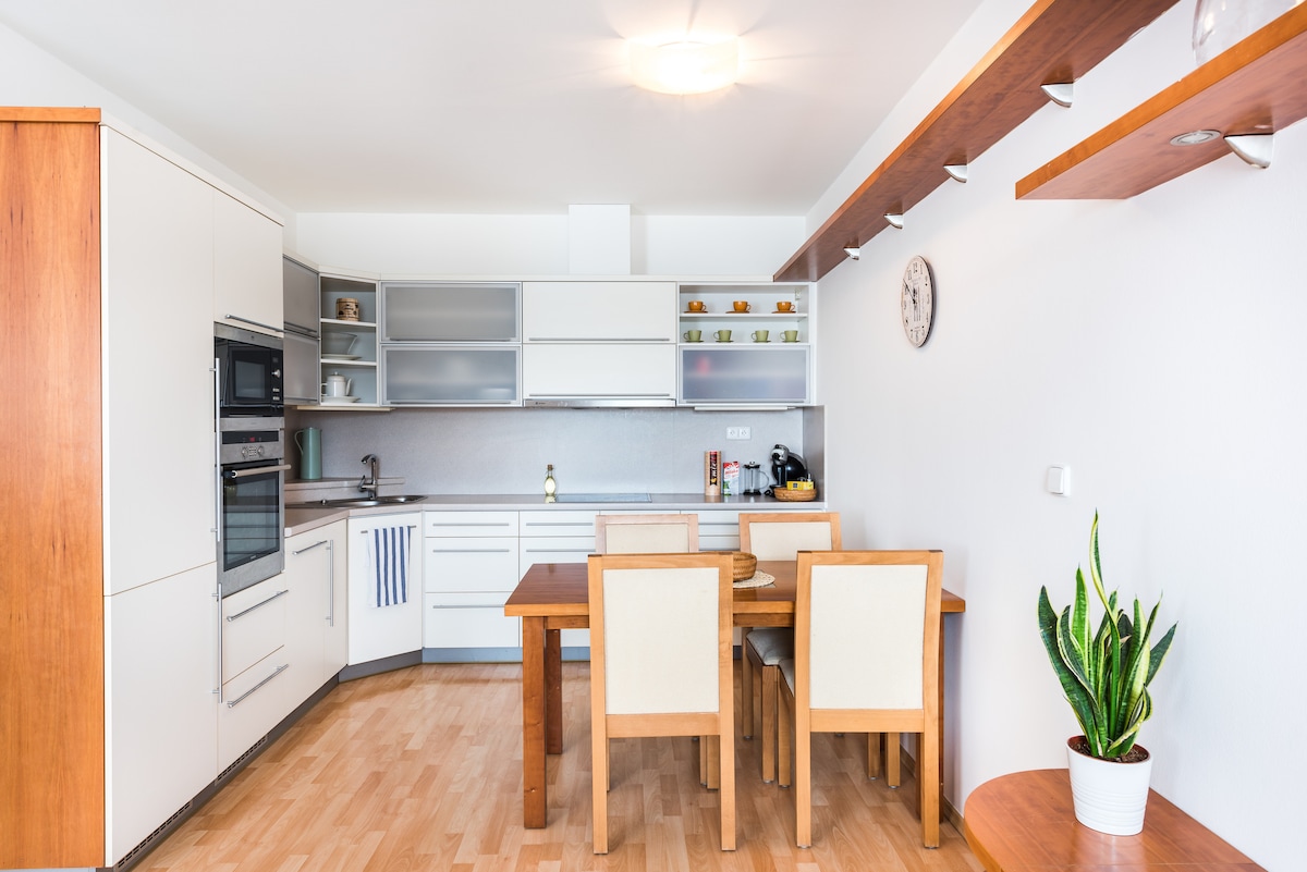 A modern kitchen and dining area is displayed, featuring sleek white cabinetry and wood accents. A dining table with four chairs is positioned nearby. Natural light fills the space, highlighting the countertop and the decorative plant placed on a shelf.