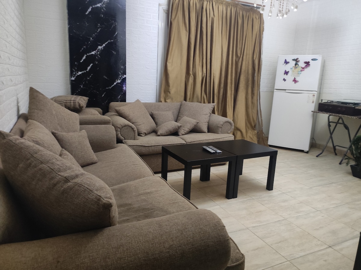 A comfortable living room features a large, plush sofa with several cushions arranged casually. A black coffee table sits in front, and a white refrigerator is visible in the background. Soft, golden curtains cover a window, providing a cozy backdrop to the space.