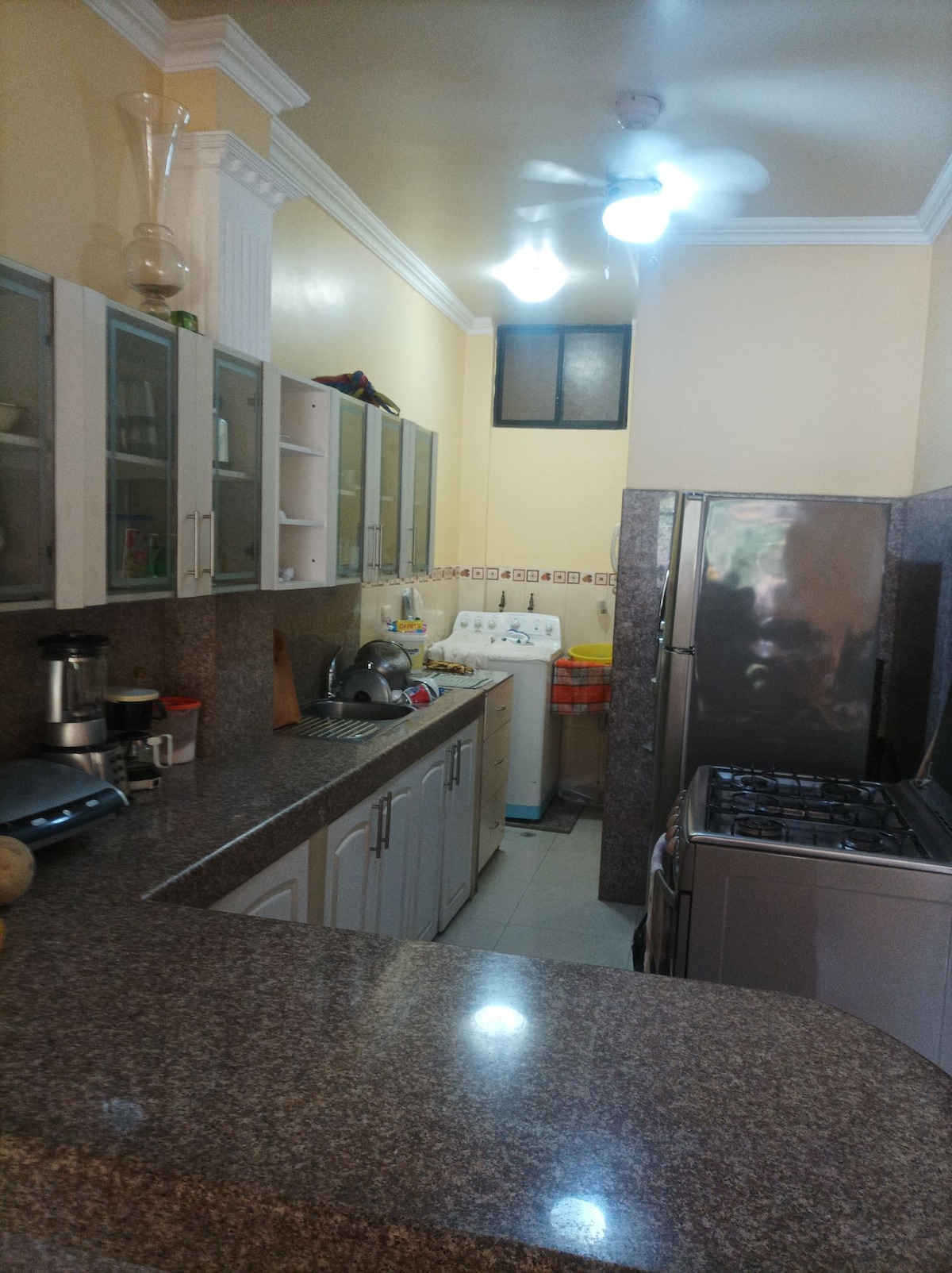 A well-organized kitchen features light-colored cabinetry and a spacious countertop. Appliances include a gas stove and refrigerator, positioned against the granite surfaces. A window allows natural light to enter, while shelves display various kitchen items, creating a functional and inviting cooking space.