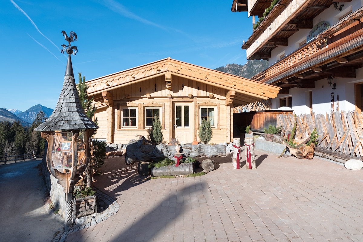 The exterior of the chalet features traditional wooden architecture with large window panes framed in natural wood. Decorative elements include a small wooden structure with a pointed roof and stone accents. Scenic mountain views are visible in the background under a clear blue sky.