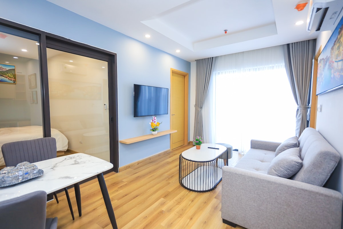 A modern living area features a comfortable sofa facing a wall-mounted television. Natural light enters through large windows, with sheer curtains gently diffusing the brightness. A round coffee table sits in the center, complemented by a dining area with a marble table and chairs.