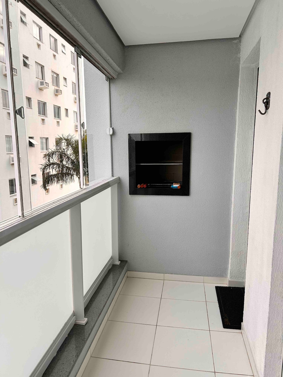 A balcony area is shown, featuring large glass windows that allow natural light to enter. A charcoal barbecue grill is mounted on the wall, while neutral-colored tiles cover the floor. A welcome mat is positioned at the entrance.