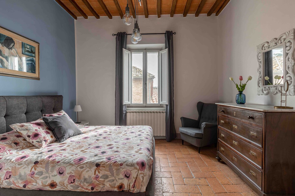 A spacious bedroom features a floral-patterned bedspread draped over a large bed. Natural light filters through a window with sheer curtains, illuminating an armchair and a wooden dresser. Decorative elements include a mirror and a framed artwork on the wall.