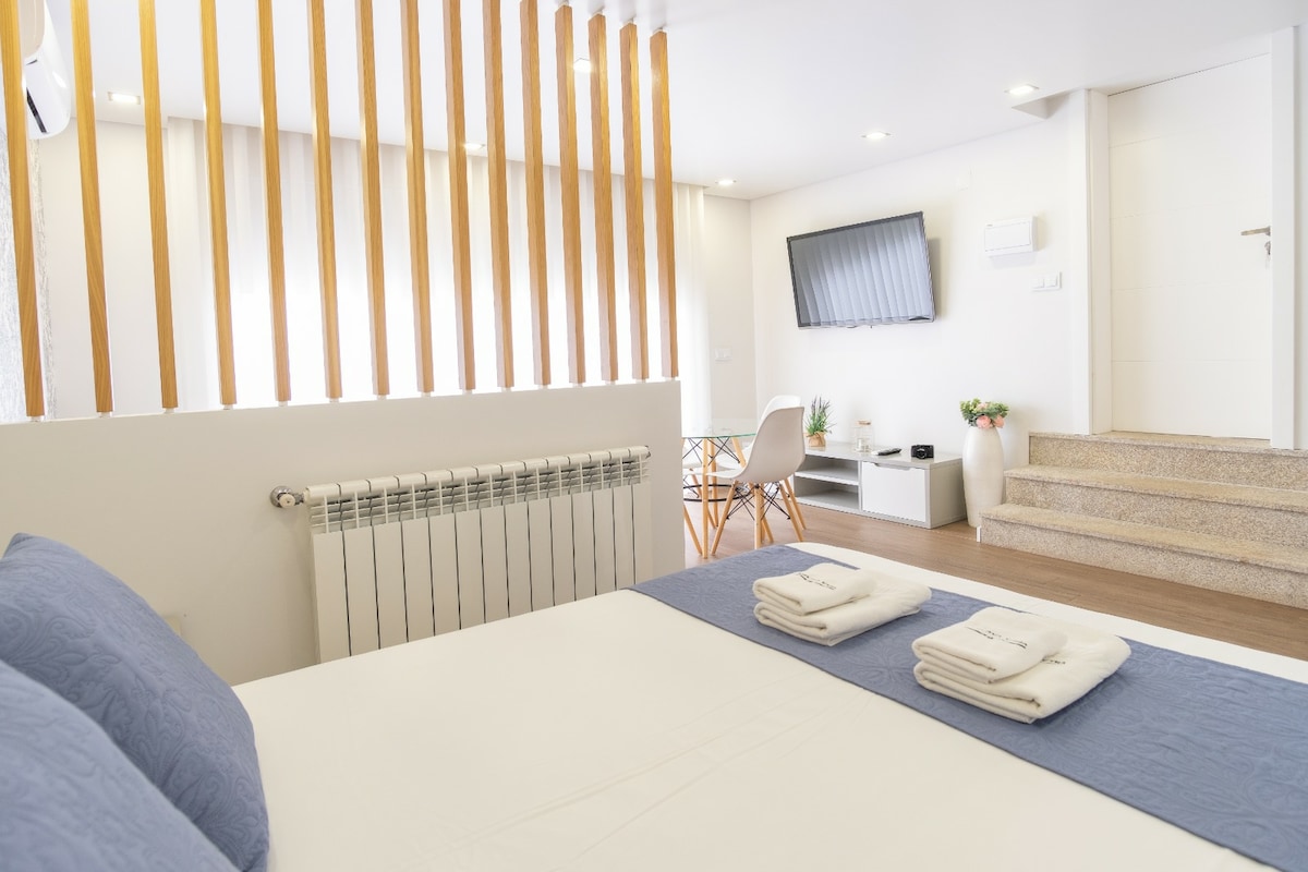 An inviting interior showcases a spacious bed with blue accents and neatly arranged towels. A modern television is mounted on the wall, and a dining area with a minimalist table and chairs is visible. Steps lead to an upper level, contributing to the room's open layout.