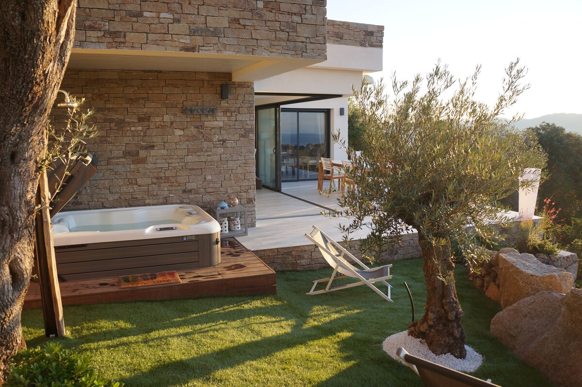 An outdoor area is showcased with a wooden deck featuring a jacuzzi and a lounge chair on a grassy surface. Natural stone walls form part of the house, with expansive glass doors leading inside, allowing for scenic views of the surrounding landscape.
