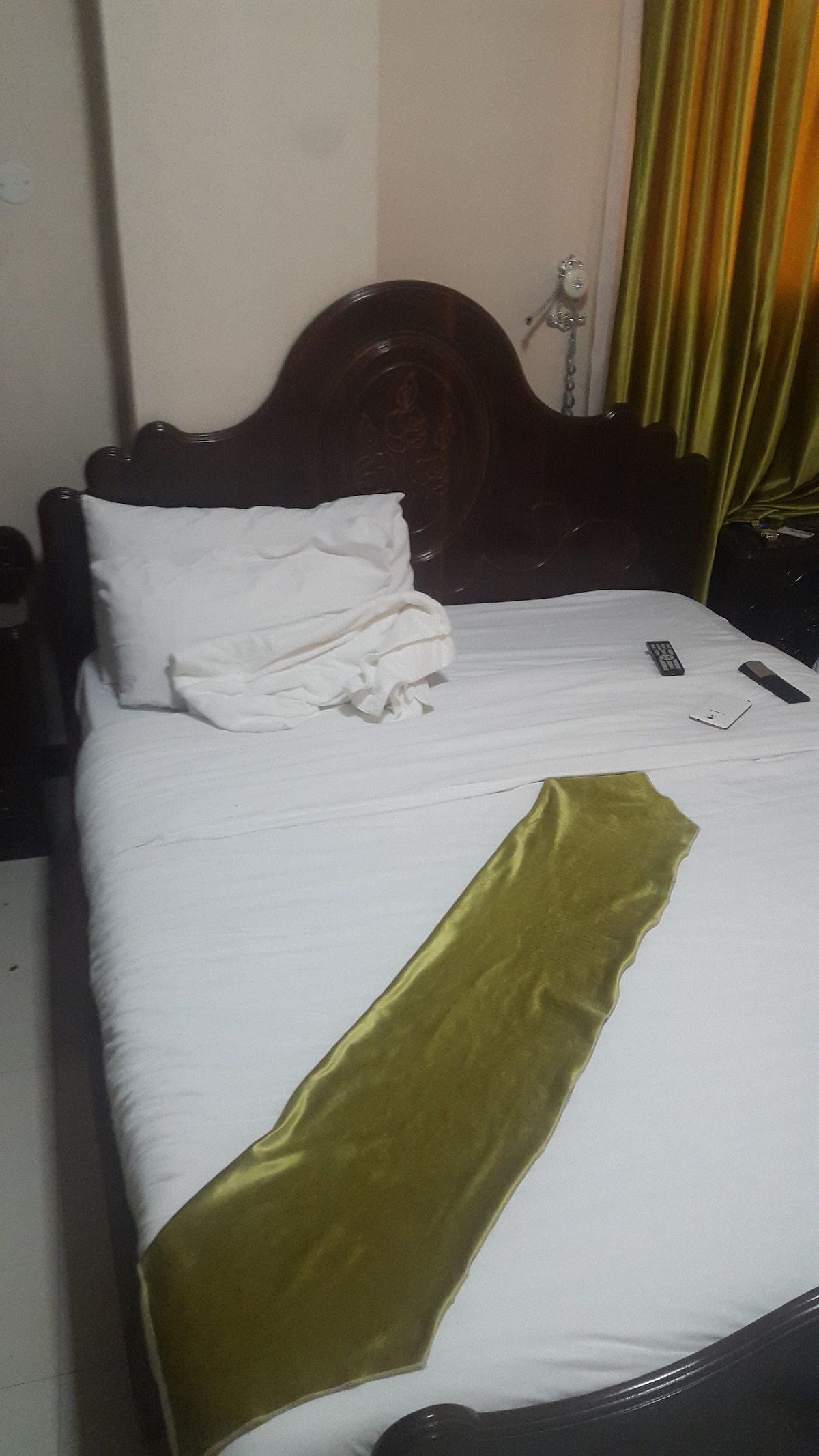 A bed with a dark wooden headboard is featured, covered in white linens. A wrinkled green throw is laid across the foot of the bed. A folded pillow sits at the head of the bed, complemented by a remote control and other small items on the side.