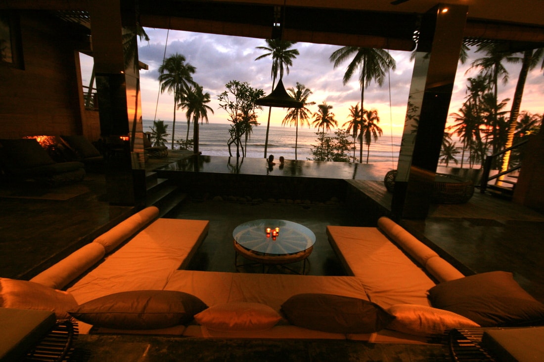 A spacious living area is framed by large glass walls, offering an unobstructed view of the ocean and palm trees. Soft seating is arranged around a circular table, with gentle lighting from candles creating a relaxing ambiance.