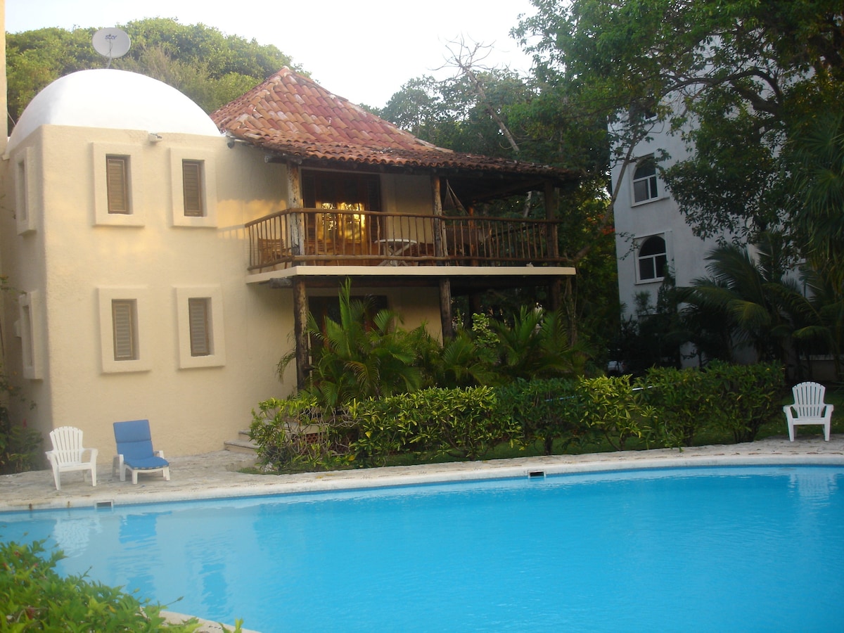 A two-story villa with a red-tiled roof and wooden balcony overlooks a large, inviting pool. The surrounding tropical gardens feature lush greenery and white lounge chairs placed near the water's edge.