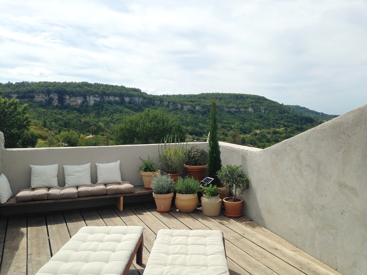 A spacious terrace features comfortable outdoor seating and assorted potted plants. The wooden floor complements the natural landscape, which includes rolling hills and greenery, creating an inviting setting for relaxation while enjoying the scenic views.
