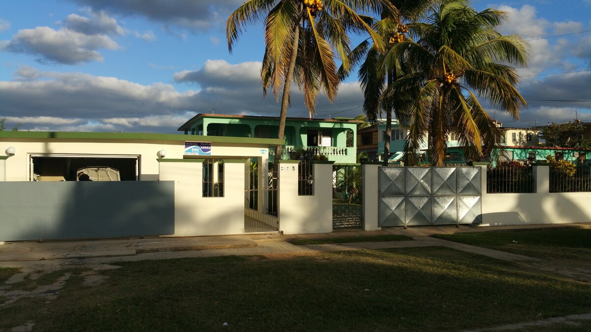 The exterior of a modern house is visible, surrounded by lush greenery and tall palm trees. A welcoming entrance features a gate with a metallic design, while light clouds drift across a blue sky, creating a tranquil setting. The well-maintained lawn adds to the inviting appearance.
