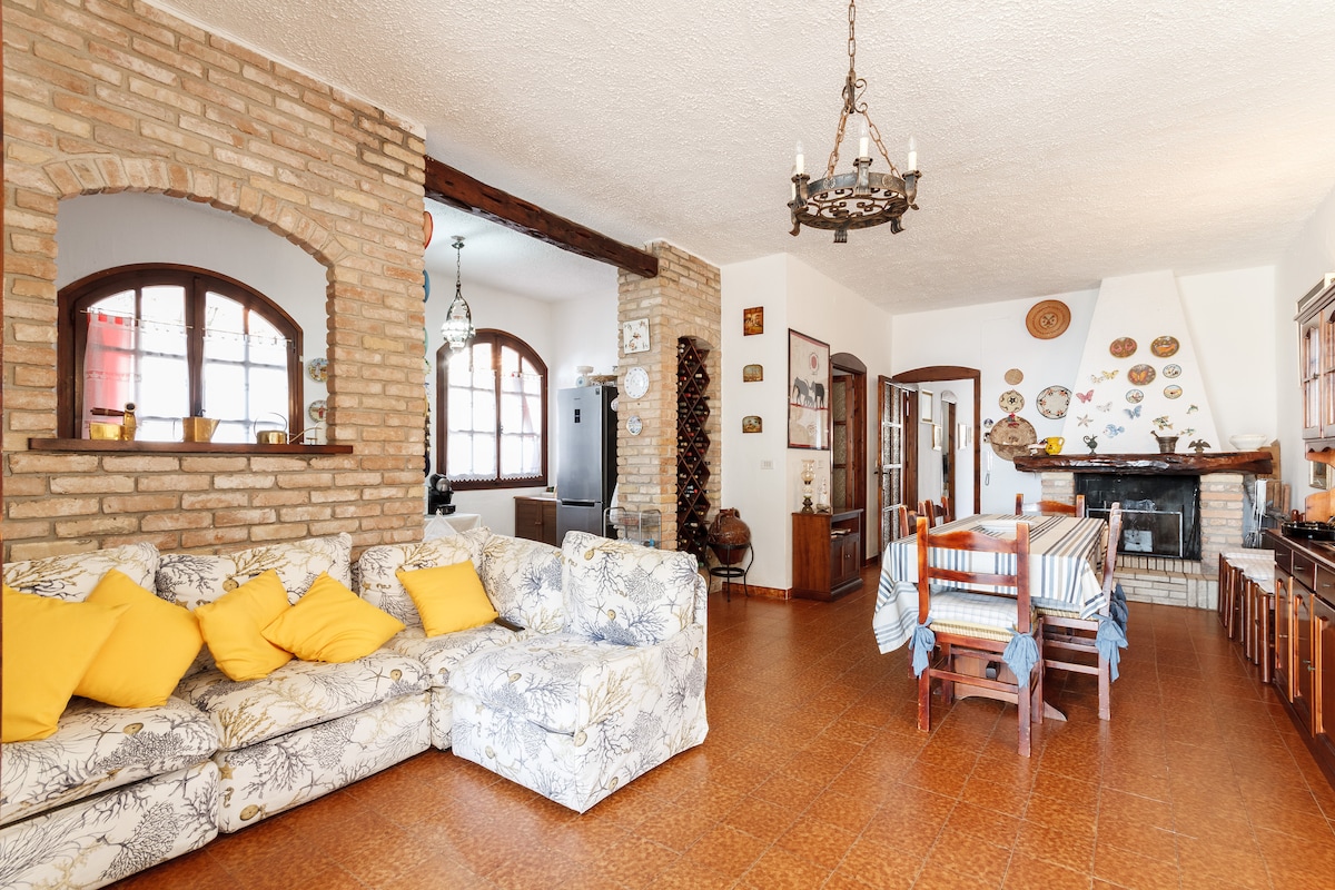 A spacious living and dining area features a large floral-patterned sofa adorned with yellow cushions. Wooden beams and brick walls lend a rustic charm. A dining table with multiple chairs sits adjacent to a fireplace, creating a welcoming environment for gatherings.