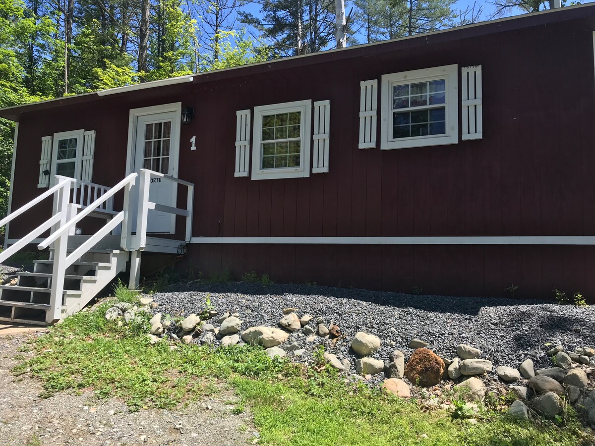 The exterior of a cozy cottage is shown, featuring red wood siding and white trim. It includes a welcoming entrance with a set of steps and a white railing. Stone landscaping surrounds the base, complemented by green grass and trees in the background.