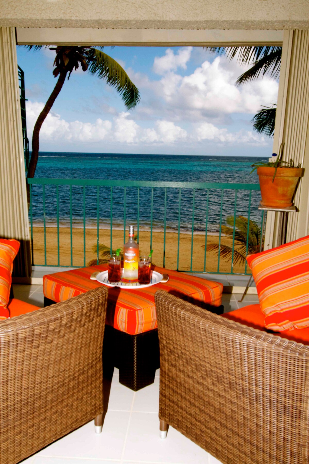 A welcoming outdoor porch is framed by a sliding glass door, offering views of the ocean and sandy beach. Two wicker chairs with orange striped cushions sit on a tiled floor, alongside a small table featuring a tray with drinks. Palm trees sway gently in the background.