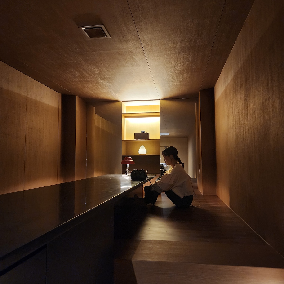 A warmly lit dining area features smooth wooden walls and a sleek, dark countertop. Subtle lighting accents the space, creating a cozy atmosphere. A person sits comfortably at the counter, surrounded by an inviting interior that emphasizes simplicity and natural materials.