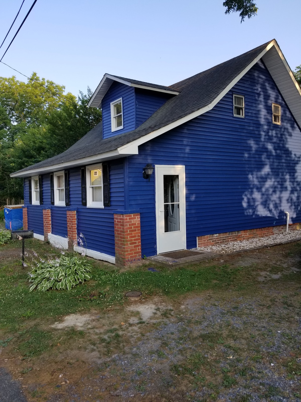 The exterior of a historic blue home is captured in this image. The house features two levels, with a gable roof and a welcoming front door. Brick accents are visible near the foundation, complemented by a neatly maintained yard and surrounding greenery.