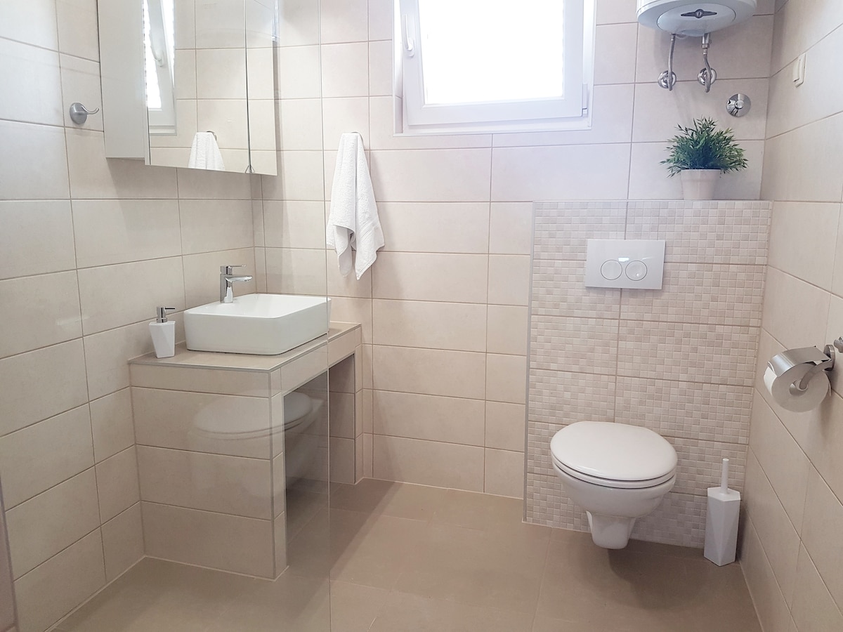 A modern bathroom features neutral tiled walls and a sleek vanity with a square sink. A large mirror is above the sink, reflecting natural light from a window. A clean toilet and a towel hang nearby, providing essential toiletries.