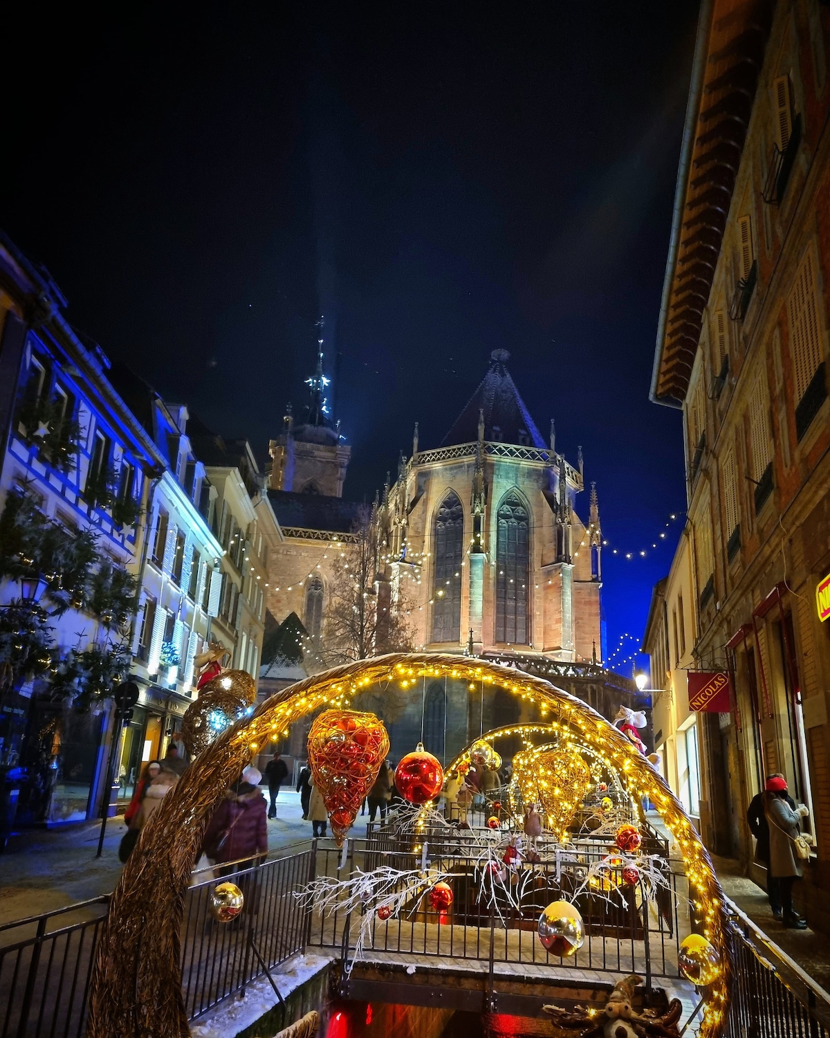 A charming street scene is illuminated at night, showcasing a historic church with intricate architecture in the background. Decorative lights adorn the trees and the pathway, enhancing the festive ambiance. The scene captures a blend of cultural heritage and seasonal celebration.