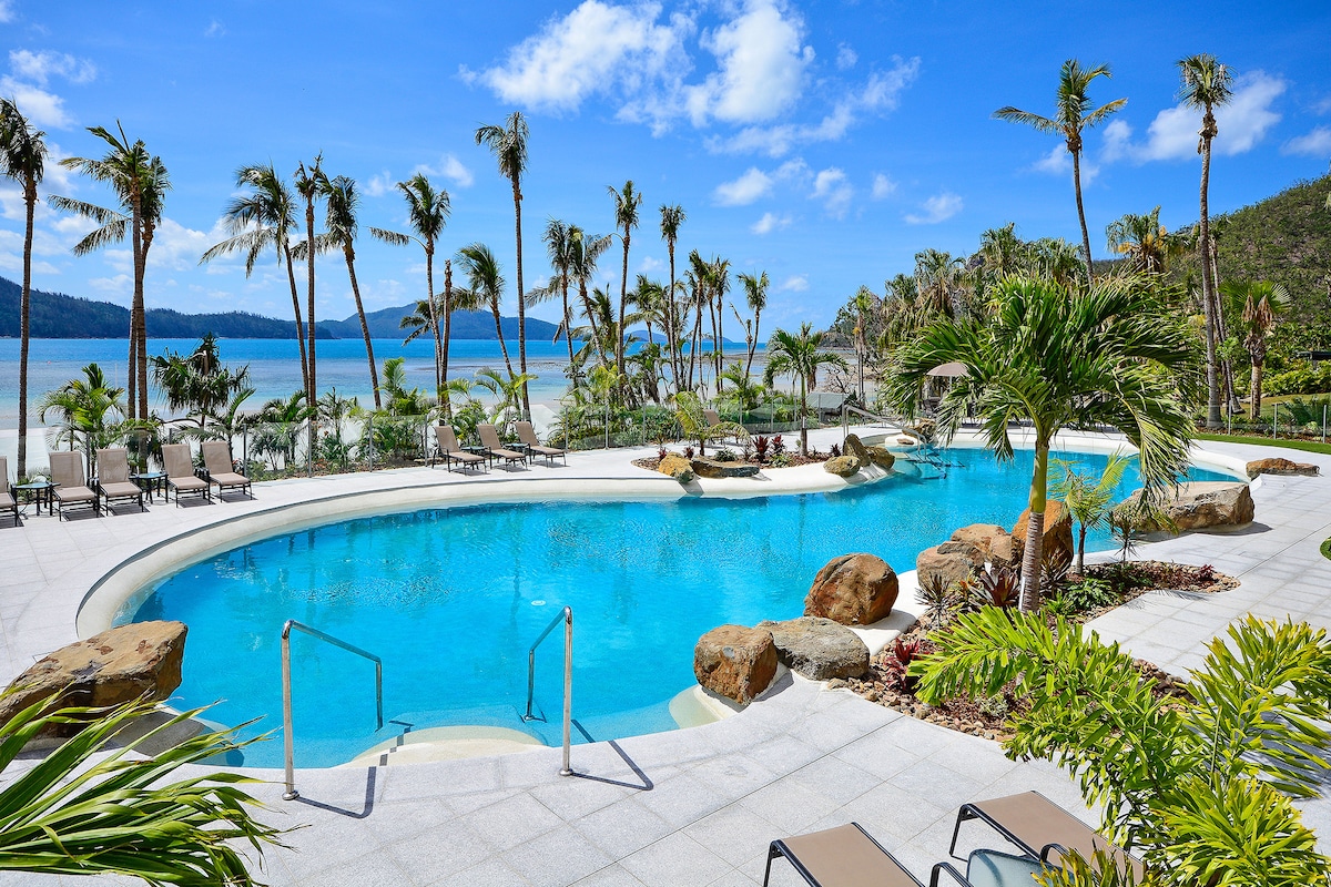 A spacious pool area is surrounded by lush tropical palm trees and natural rock features. Crystal-clear water sparkles under the sun, with poolside loungers arranged neatly nearby. A stunning view of the beach and distant hills can be seen in the background, creating a serene atmosphere.