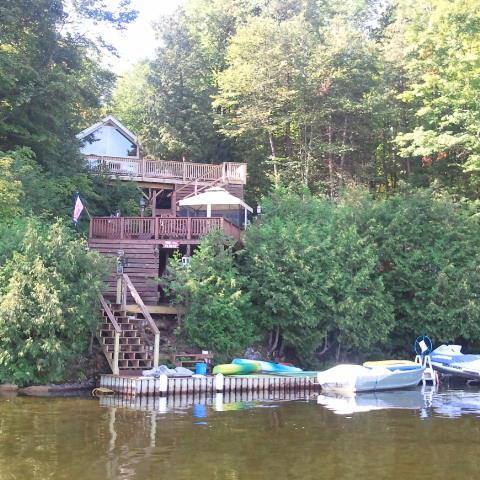 The cottage, situated on the East Shore of Lake Bonaparte, is shown with upper and lower decks, surrounded by lush greenery. A dock is visible, accommodating various watercrafts, while steps lead from the water to the decks, creating easy access for guests.