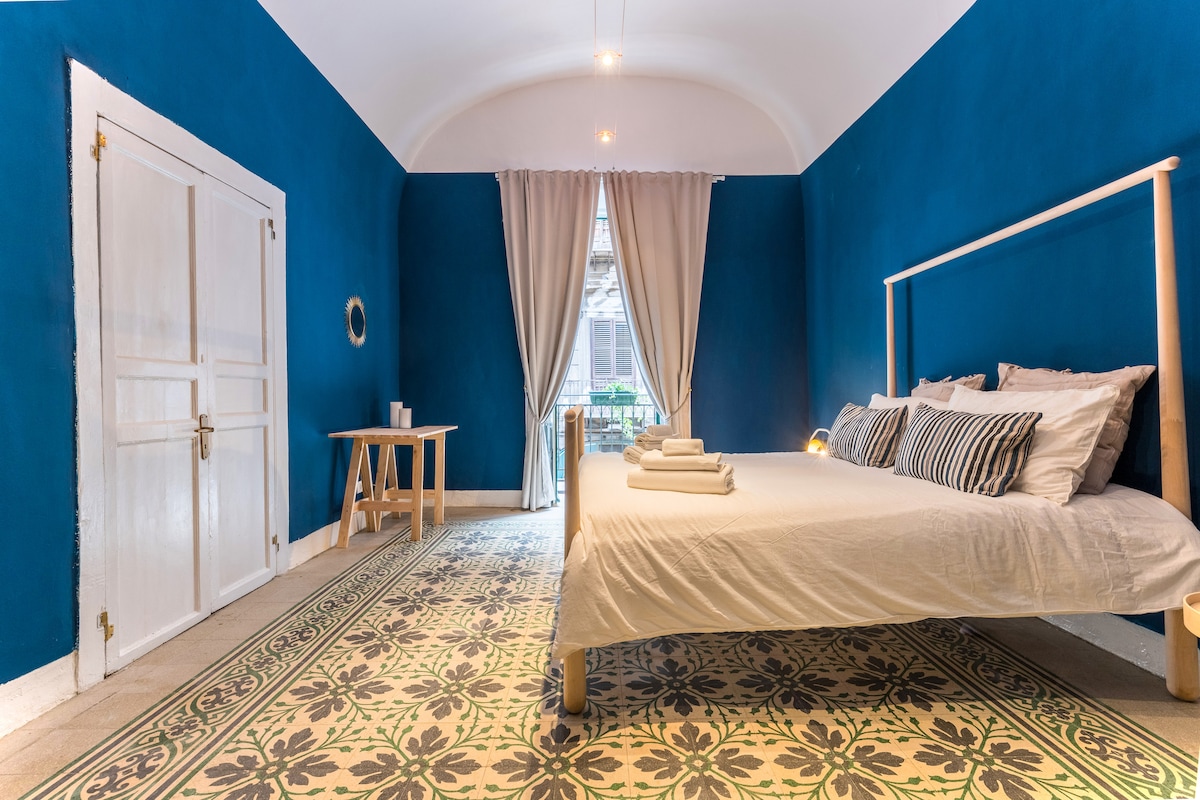 A spacious bedroom features bold blue walls that create a striking backdrop. A large bed is dressed in white linen with neatly arranged towels. An ornate area rug covers the floor, and natural light enters through a balcony window, complemented by soft curtains.