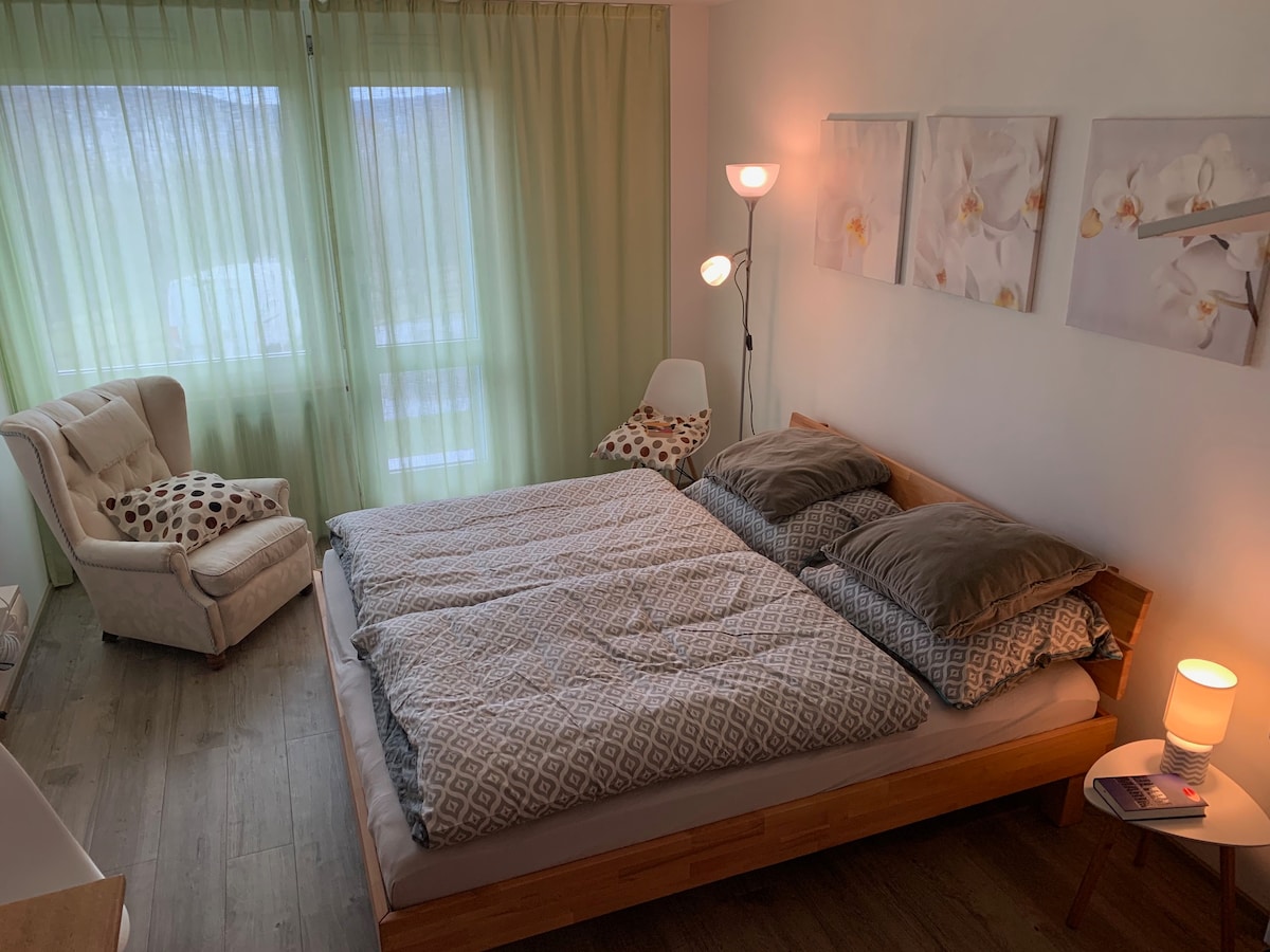 A bright sleeping area is featured with a spacious wooden bed covered with patterned bedding. Two large windows dressed in light green curtains allow natural light to fill the space. A cozy armchair and floor lamp complement the decor, which includes floral wall art.