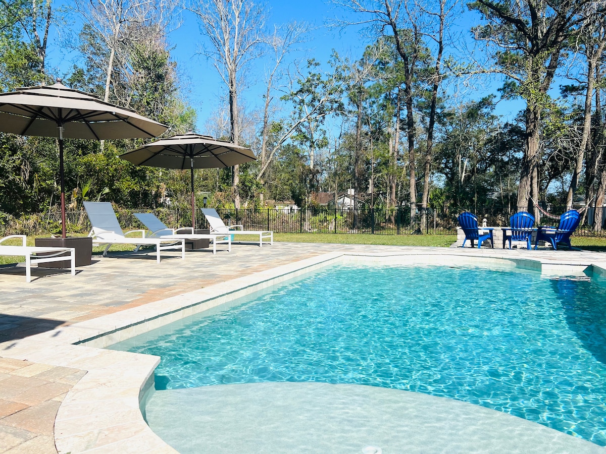 A private swimming pool is surrounded by a spacious patio, featuring white lounge chairs and two large umbrellas for shade. Comfortable blue seating is positioned nearby, offering a relaxed outdoor gathering space. Lush greenery forms the backdrop, enhancing the tranquil setting.